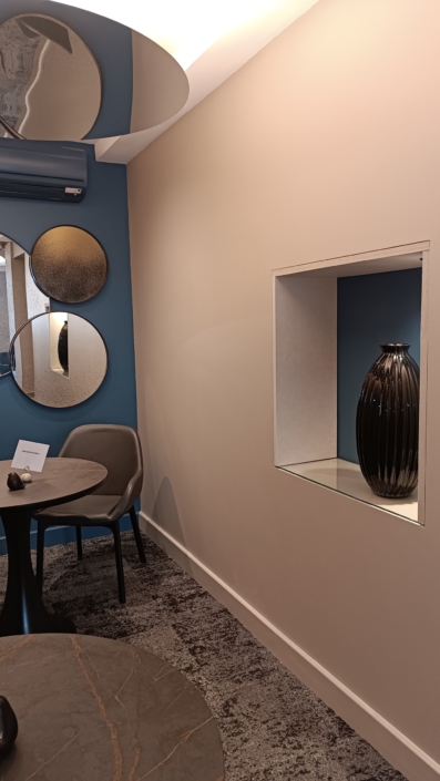 Cette pièce moderne dispose d'un agencement de miroirs circulaires, d'une petite table avec une chaise et d'une niche affichant un vase sombre, complétés par d'élégants panneaux acoustiques suspendus pour un style et une insonorisation améliorés.
