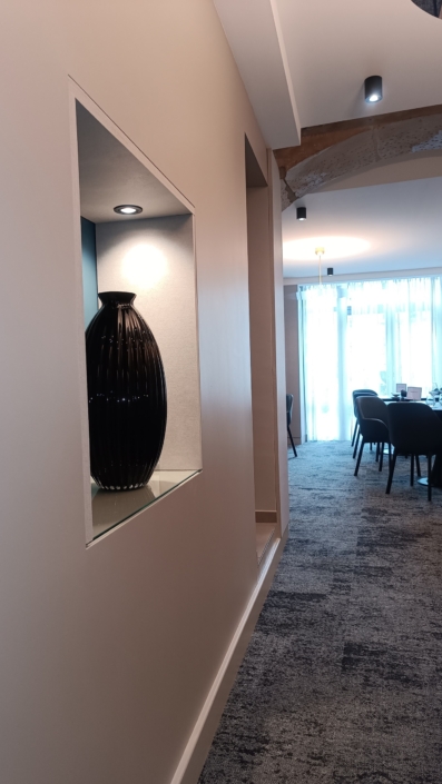 Un couloir épuré est orné d'un grand vase noir dans une niche, menant à une salle à manger ornée d'élégantes chaises sombres et de grandes fenêtres. Des panneaux acoustiques suspendus muraux rehaussent subtilement l'ambiance tout en offrant une qualité sonore supérieure.