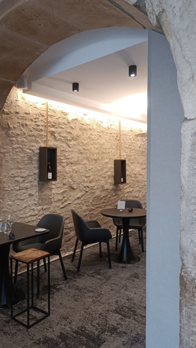Intérieur de restaurant chaleureux avec murs en pierre, chaises noires modernes, tables rondes et éclairage tamisé. Le design est complété par des panneaux acoustiques élégants au plafond pour améliorer l'ambiance.