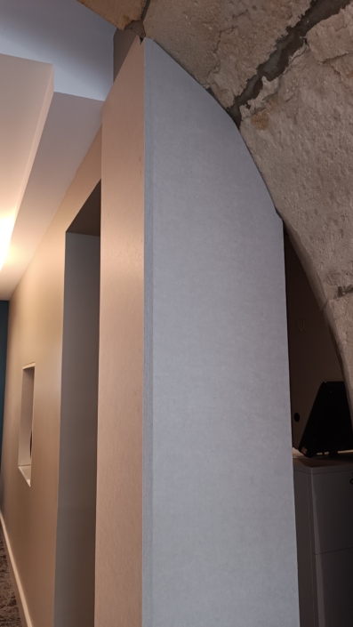 Un mur moderne croise une arche en pierre, soulignant le contraste entre les styles architecturaux anciens et nouveaux. Suspendus au plafond, des panneaux acoustiques épurés ajoutent une touche de style contemporain tout en améliorant la qualité sonore de l'espace.