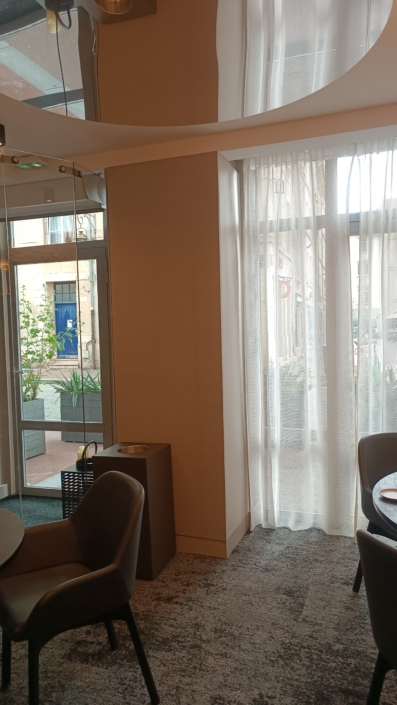 Coin restaurant cosy avec chaises, table et rideaux transparents près d'une fenêtre laissant entrer la lumière naturelle, complété par d'élégants panneaux acoustiques suspendus doucement d'en haut.