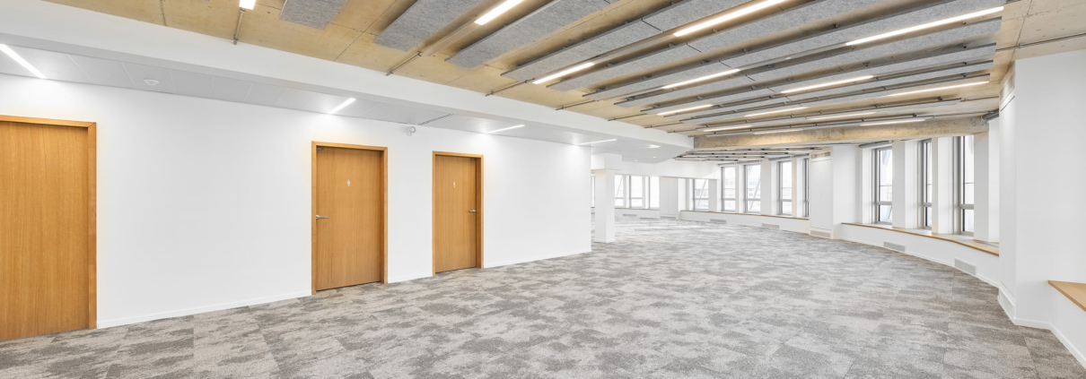 Large empty room with gray carpet, wooden doors and a ceiling with acoustic panels reminiscent of an acoustic gymnasium, with integrated lights.