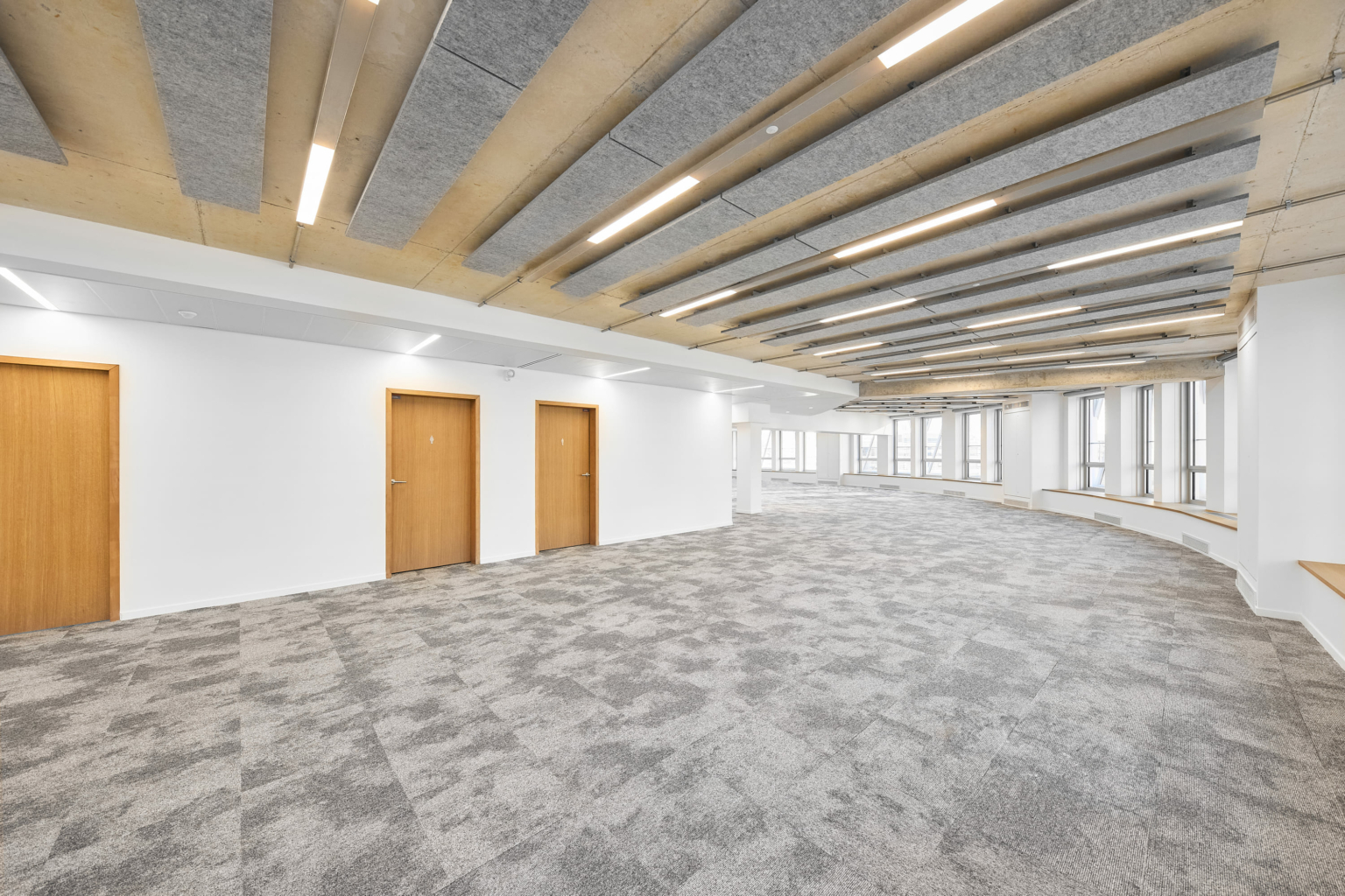Grande salle vide avec moquette grise, portes en bois et plafond doté de panneaux acoustiques rappelant un gymnase acoustique, avec lumières intégrées.