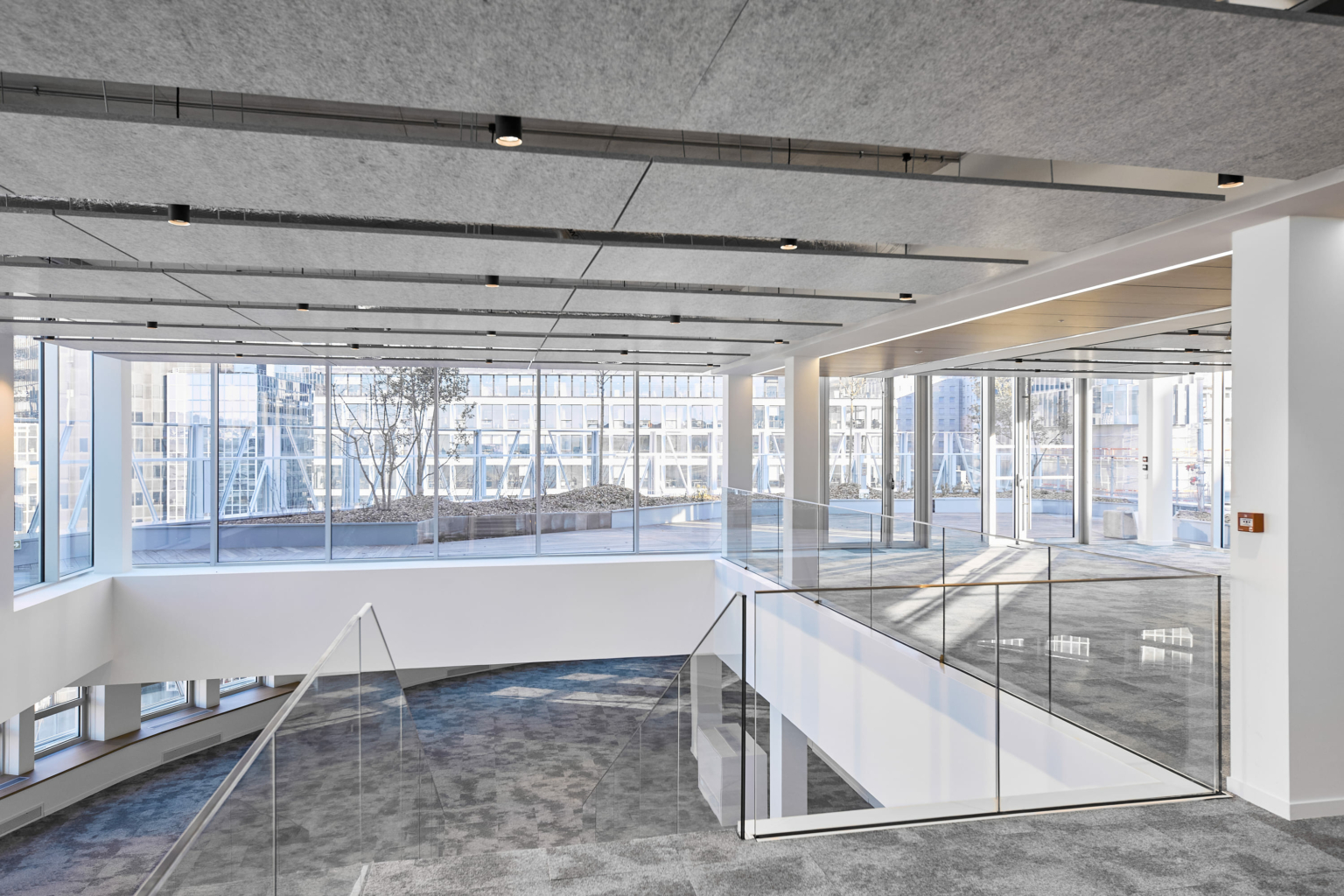 Le hall d'entrée moderne, doté d'un design insonorisant avec de grandes fenêtres, des balustrades en verre et un sol gris, offre une vue sereine sur le paysage urbain animé.