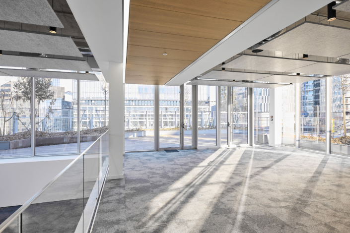 Un espace de bureau moderne et vide avec de grandes fenêtres, un tapis gris et des panneaux de plafond en bois offre une atmosphère sereine rappelant un gymnase acoustique, tout en donnant sur un paysage urbain animé.