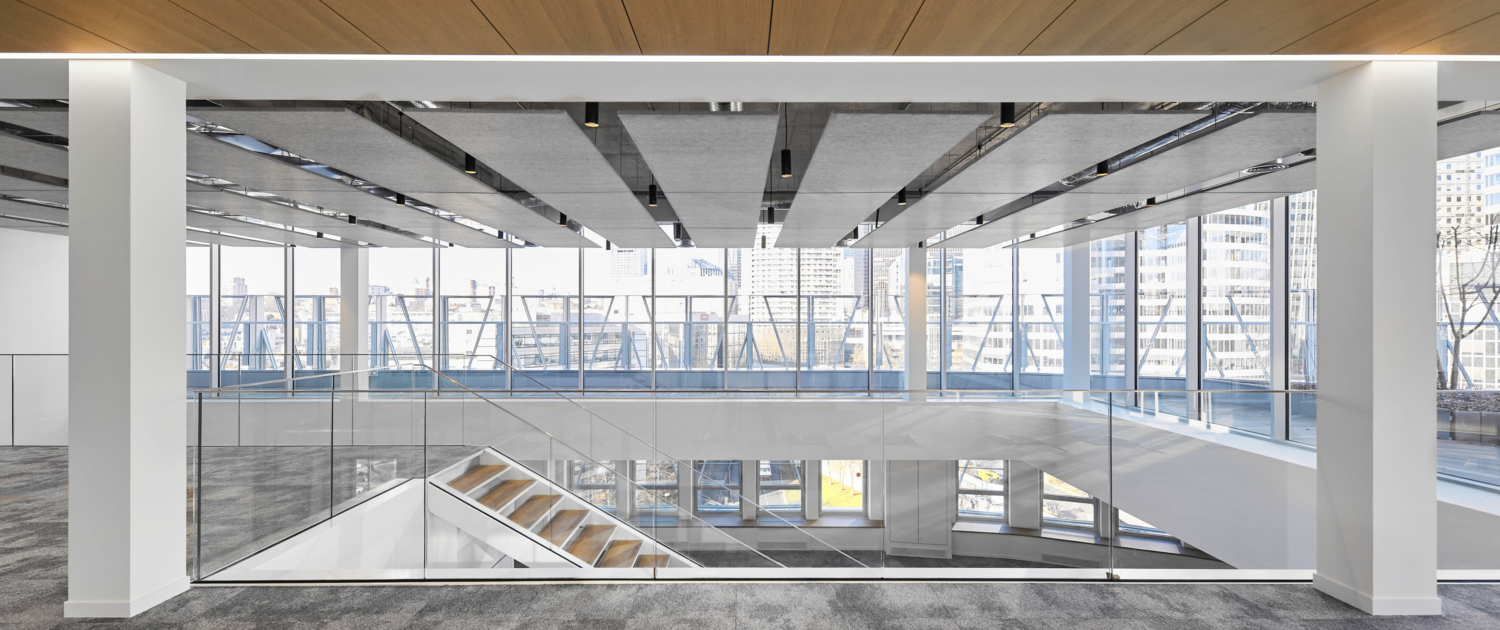 Espace de bureau moderne avec murs en verre et escalier ouvert, doté de grandes fenêtres offrant une vue sur la ville et des améliorations acoustiques semblables à celles d'un gymnase pour minimiser le bruit.