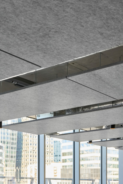 Un plafond moderne avec des panneaux gris et des accents métalliques, conçu acoustiquement comme un gymnase, est placé contre de grandes fenêtres présentant un paysage urbain animé.