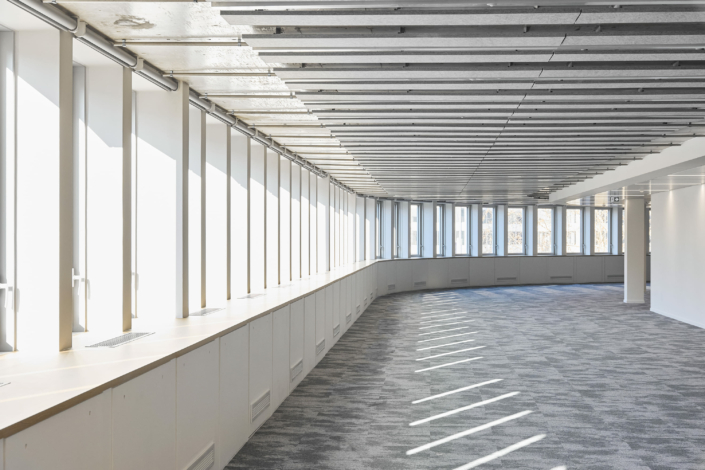 L'espace de bureau tout en longueur et en courbes, rappelant un gymnase acoustique, est doté de grandes fenêtres et d'un sol recouvert de moquette. Des poutres suspendues complètent le design tandis qu'une lumière naturelle éclatante inonde la pièce.
