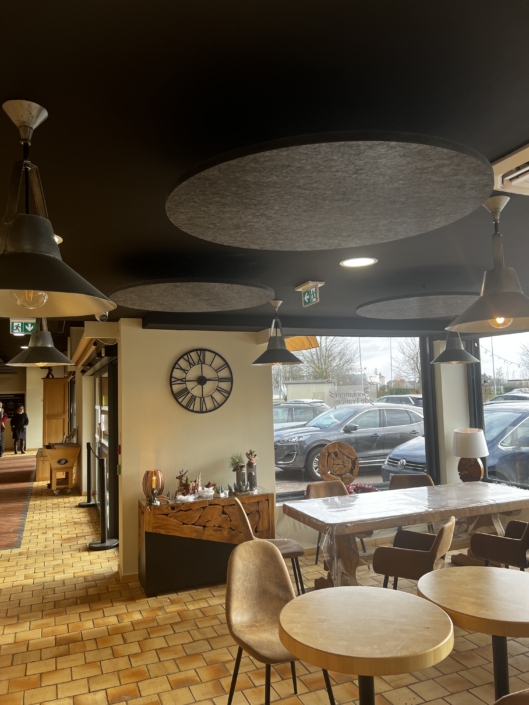 Intérieur de café avec tables rondes et décor en bois plein de charme, rehaussé par l'ambiance d'un restaurant acoustique. Une grande horloge orne le mur, tandis que les fenêtres offrent une vue sur les voitures stationnées à l'extérieur.