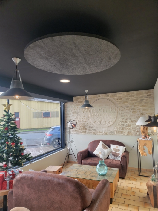 Coin salon cosy avec chaises marron, table en bois et sapin de Noël dans une pièce aux grandes fenêtres et murs en pierre, rappelant le décor d'un restaurant acoustique.