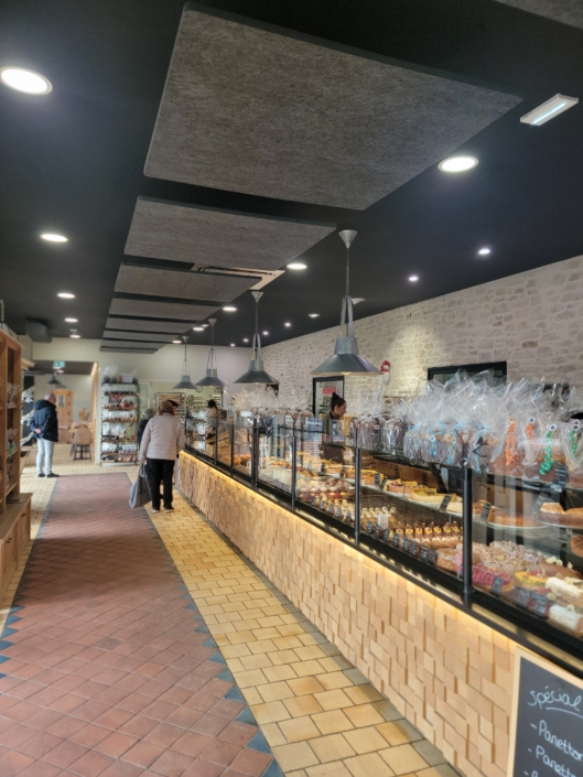 L'intérieur d'une boulangerie avec un long comptoir de présentation rempli de pâtisseries et de luminaires suspendus, rappelle un restaurant acoustique. Les gens flânent, captivés par l'ambiance accueillante et les arômes délicieux.
