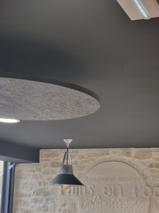 Plafonnier avec un panneau rond gris au-dessus et un mur en pierre avec un texte pâle en arrière-plan, rappelant une ambiance de restaurant acoustique.