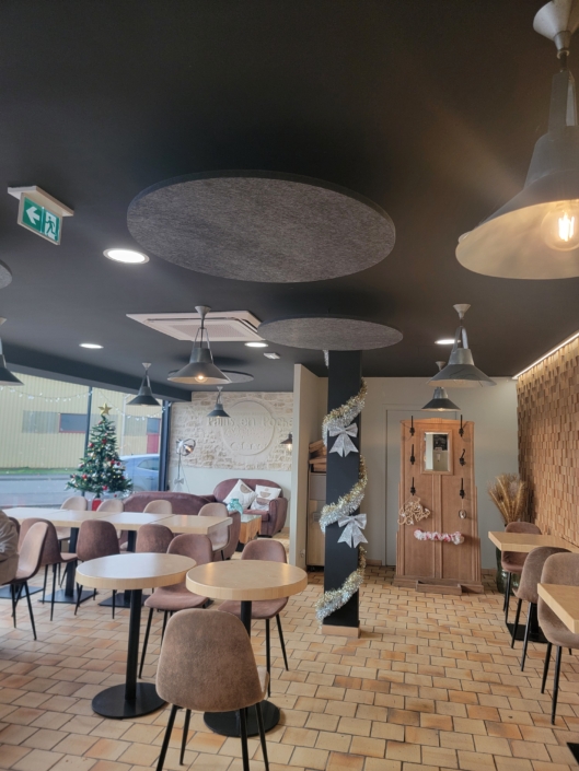 L'intérieur confortable d'un café avec des tables rondes, des chaises marron, un sapin de Noël et des lumières suspendues décoratives évoque l'ambiance chaleureuse d'un restaurant acoustique.