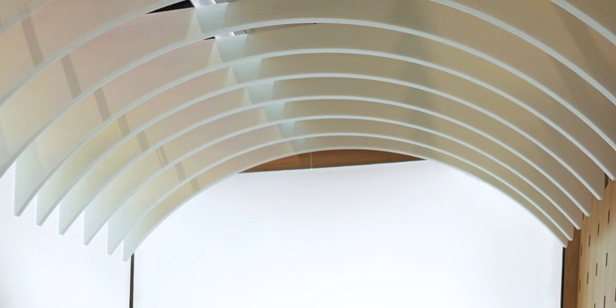 Panneaux de plafond blancs incurvés disposés en lignes parallèles sur un mur blanc dans un espace intérieur moderne.