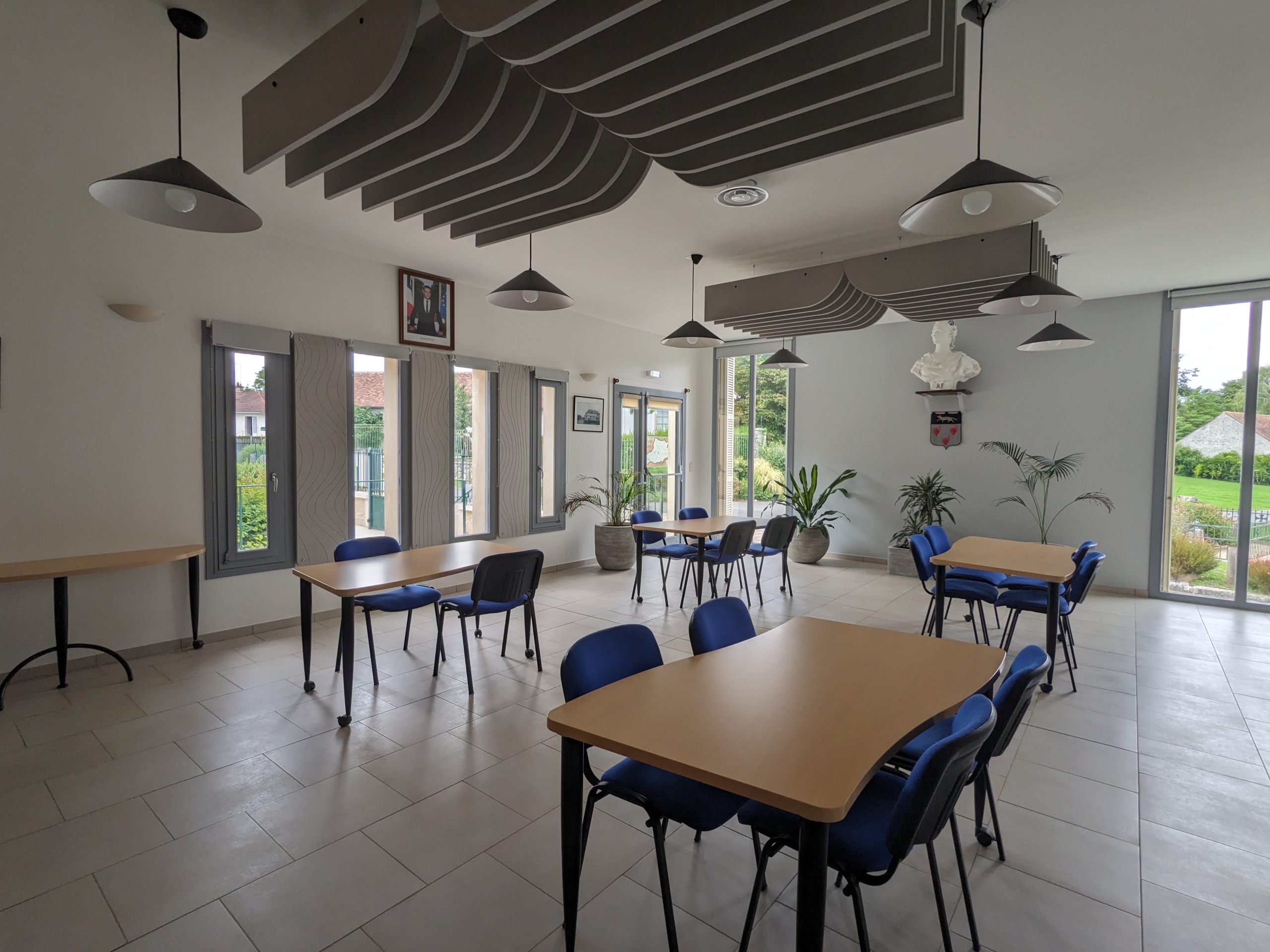 Une salle vide et lumineuse avec des tables et des chaises bleues, de grandes fenêtres, des plantes et des plafonniers modernes.
