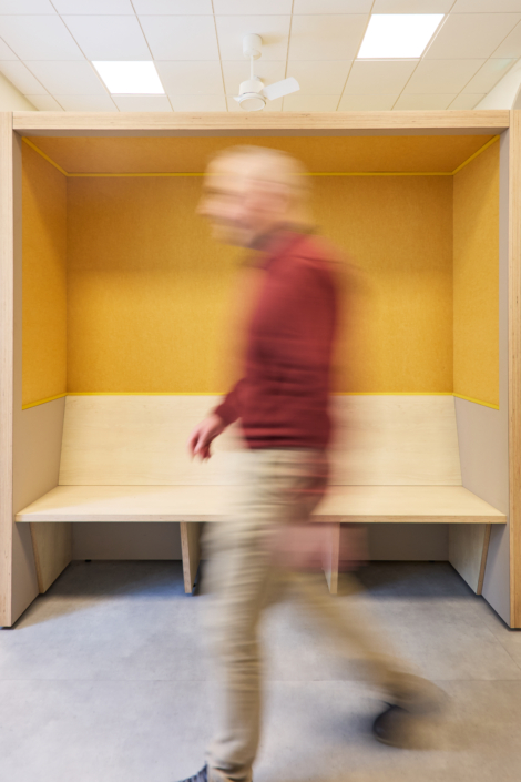 Une personne en mouvement passe devant un banc en bois doté d'un dossier jaune, dans une salle moderne conçue pour optimiser l'acoustique d'un gymnase.