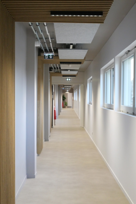 Hall d'entrée moderne avec un sol beige, des murs blancs, de grandes fenêtres à droite et des éléments décoratifs en bois au plafond, conçus pour offrir une qualité sonore inspirée de celle d'un gymnase.