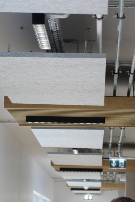 Plafond composé de panneaux acoustiques de gymnase, de tuyaux apparents et de luminaires LED dans un couloir moderne.
