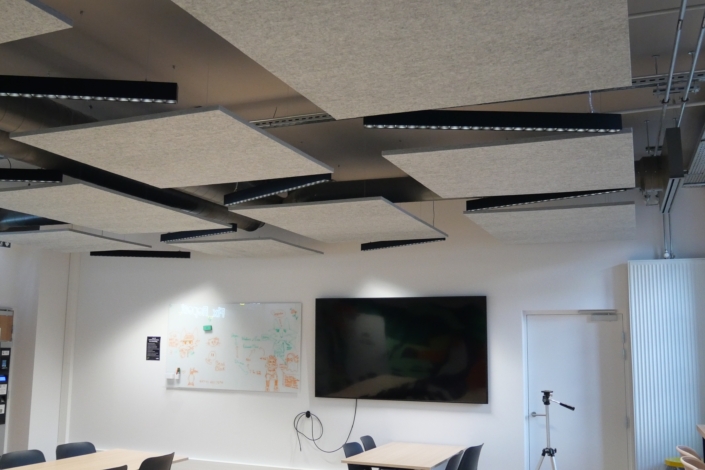 Des panneaux acoustiques et des luminaires sont suspendus au plafond de cette salle de classe moderne — idéale pour répondre aux besoins acoustiques d'un gymnase —, équipée de tables, d'un téléviseur et d'un tableau blanc.
