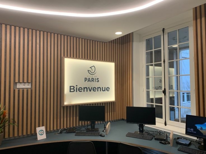 Una zona reception con pareti in legno, arredata nello stile di una palestra acustica, dotata di computer e con un cartello recante la scritta "City of Paris Welcome" vicino a una finestra.