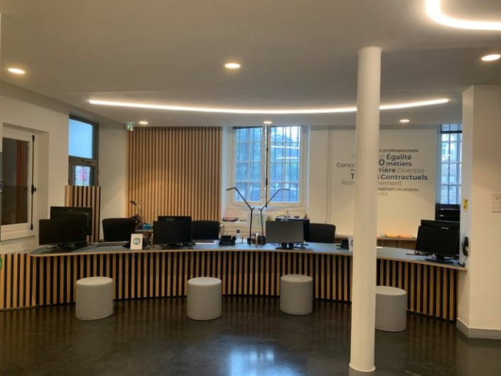 Una moderna area reception per ufficio con pannelli in legno, diverse scrivanie, computer e sgabelli rotondi grigi disposti di fronte. Questo spazio, progettato con cura, trae ispirazione dalle soluzioni acustiche utilizzate nelle palestre per creare un'atmosfera calma e accogliente.