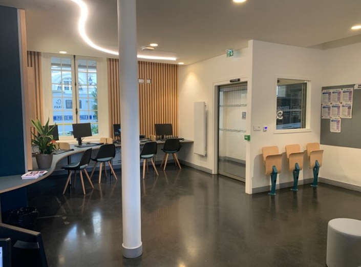 Moderna sala d'attesa con sedie, scrivanie, una pianta e una finestra; pareti in legno chiaro e bianco, design pulito ispirato ai principi acustici delle palestre per maggiore comfort e tranquillità.