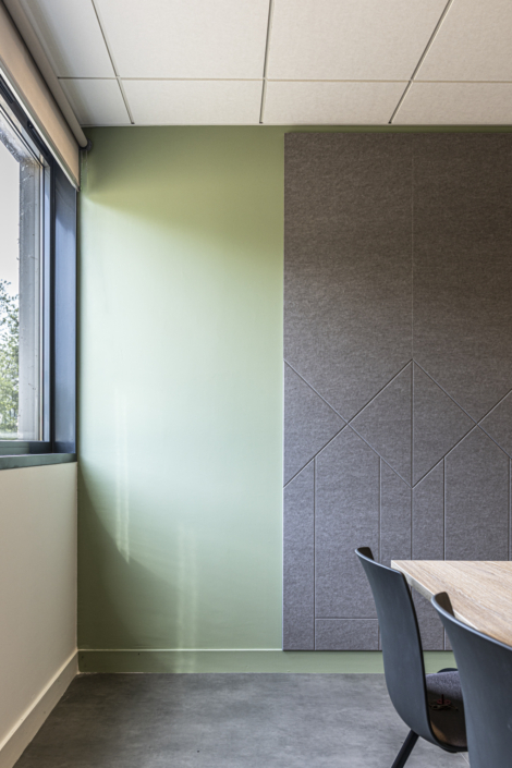 Angolo ufficio moderno con parete verde, pannello grigio, finestra e vista parziale di sedie e tavolo, caratterizzato da elementi di design ispirati all'acustica delle palestre per una migliore qualità del suono e un maggiore comfort.