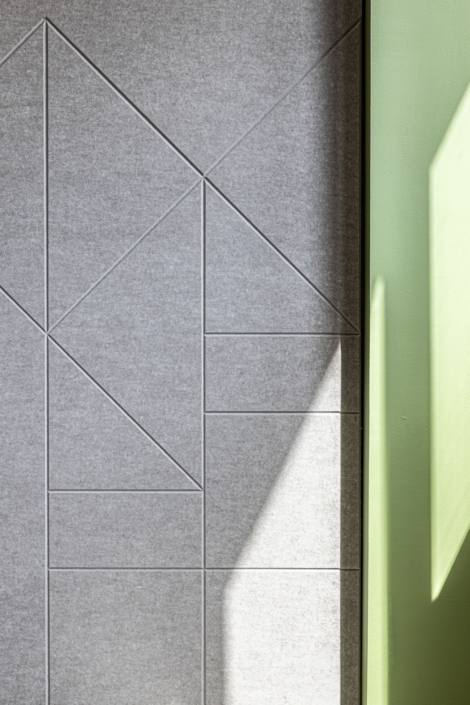 Piastrelle geometriche grigie si affiancano a una parete verde, mentre la luce del sole proietta un'ombra triangolare su entrambe le superfici, creando uno stile moderno che ricorda il design di una palestra "Acoustic".