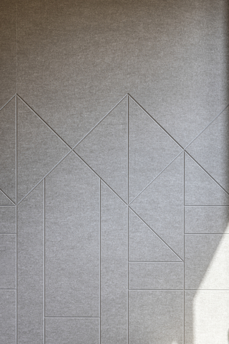 Parete grigia con superficie ruvida, decorata con linee geometriche che formano sagome astratte di montagne, progettata per migliorare l'acustica della palestra; un raggio di sole nell'angolo in basso a destra.