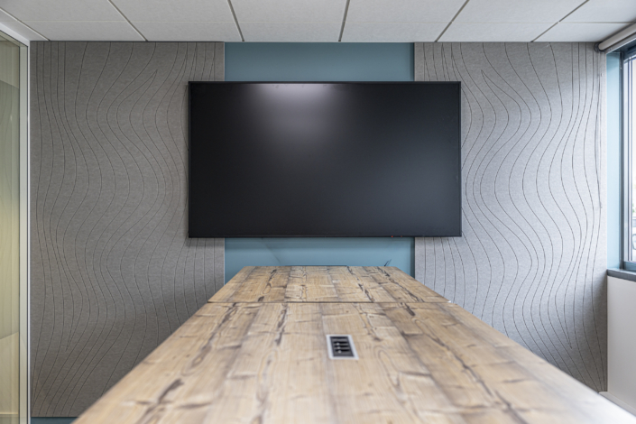 Un grande schermo televisivo montato a parete dietro un tavolo da conferenza in legno, in una moderna sala riunioni progettata con caratteristiche acustiche "da palestra" per una qualità del suono ottimale.