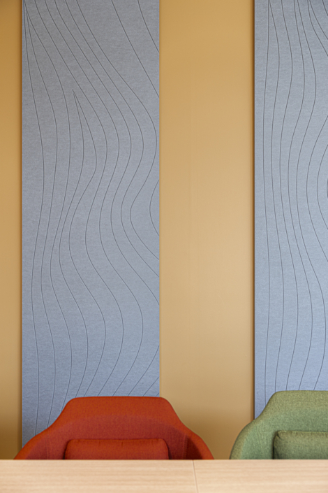 Due sedie colorate e un tavolo di legno, posizionati davanti a pareti gialle decorate con pannelli blu dai motivi ondulati, creano uno spazio vivace che richiama in modo discreto gli elementi di design della palestra "Acoustic".