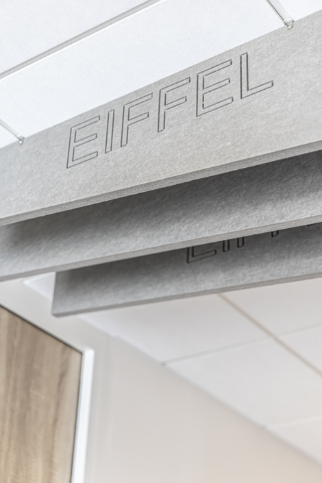 Pannello a soffitto grigio con la scritta "EIFFEL" intagliata, progettato per ottimizzare le prestazioni acustiche della palestra, sospeso in un moderno controsoffitto rivestito di piastrelle bianche.