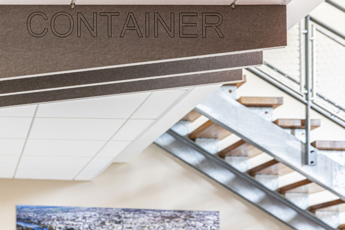 Un cartello marrone con la scritta "CONTAINER" in lettere maiuscole, posto sopra una moderna scala interna, ricorda il design pulito di una palestra acustica.