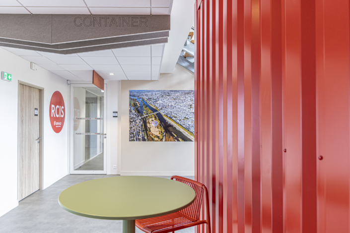 Spazio ufficio moderno con una parete rossa, un tavolo rotondo verde, una panca e una fotografia aerea appesa alla parete bianca, caratterizzato da elementi di design ispirati all'acustica delle palestre per una migliore qualità del suono.