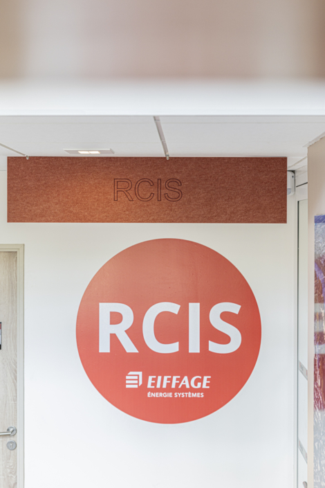 Un cerchio rosso con le scritte "RCIS" e "Eiffage Énergie Systèmes" in bianco su una parete bianca, vicino a una porta della palestra insonorizzata.