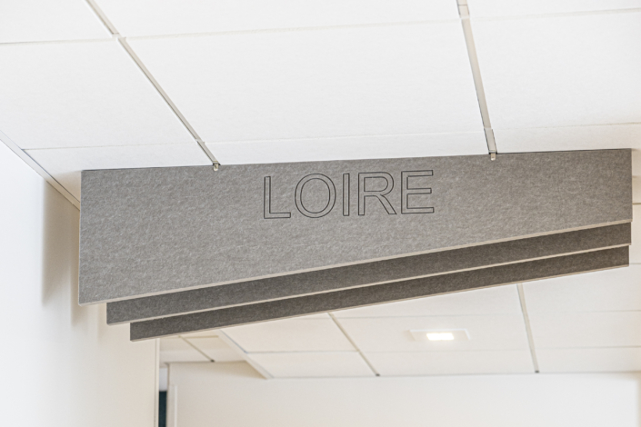 Un'insegna grigia a soffitto con la scritta "LOIRE" in lettere nere, sospesa in un corridoio con controsoffitti bianchi, che ricordano quelli che si trovano spesso nelle palestre con una buona acustica.