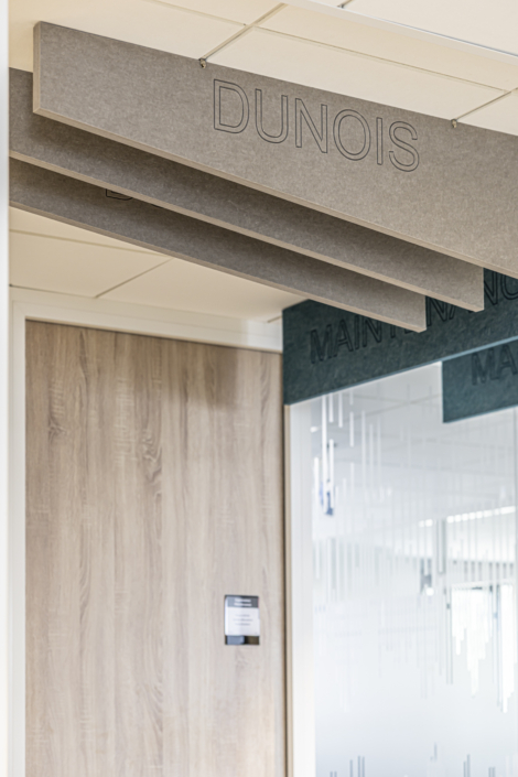 Pannelli a soffitto con la scritta "DUNOIS" sopra una porta in legno e una parete in vetro decorata con linee incise, in uno spazio ufficio progettato secondo gli standard acustici delle palestre per un assorbimento acustico ottimale.