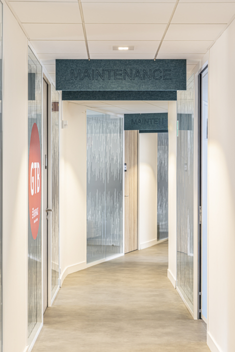 Un corridoio moderno con porte a vetri e un cartello "MANUTENZIONE" sospeso al soffitto, progettato secondo i principi acustici delle palestre per ottimizzare l'isolamento acustico.