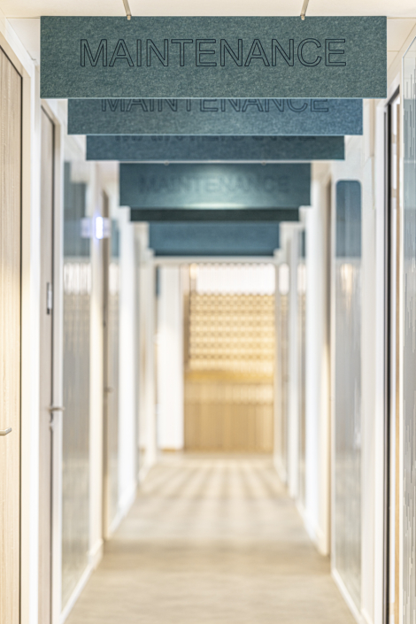 Un corridoio dove diversi pannelli blu con la scritta "MANUTENZIONE" sono sospesi al soffitto, con porte su entrambi i lati, che richiamano le linee pulite e il design funzionale tipici di una palestra acustica.