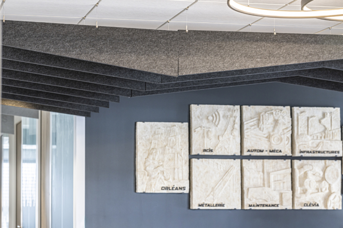 Sei targhe di pietra scolpite con iscrizioni sono fissate a una parete scura, sotto pannelli grigi del soffitto disposti ad angolo, che ricordano il design raffinato che si trova spesso in una palestra acustica.