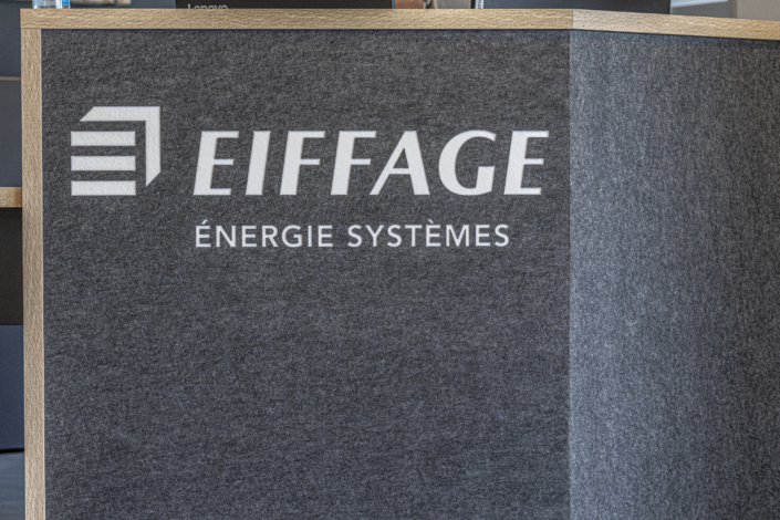 Logo di Eiffage Énergie Systèmes su un pannello murale grigio con superficie ruvida, bordato da modanature in legno, progettato per migliorare l'acustica della palestra.