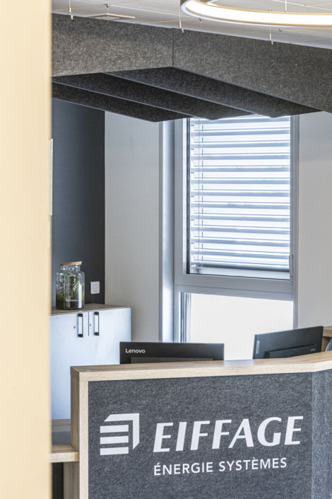 Moderna area reception degli uffici, con il logo di Eiffage Énergie Systèmes, schermi per computer e tende, progettata secondo i principi dell'acustica delle palestre per migliorare la qualità del suono e il comfort.