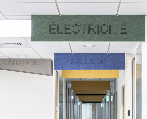 Cartelli colorati appesi al soffitto di un corridoio, con le scritte "Elettricità", "Studio medico" e "Contenitore", creano un'atmosfera dinamica che ricorda una palestra acustica.