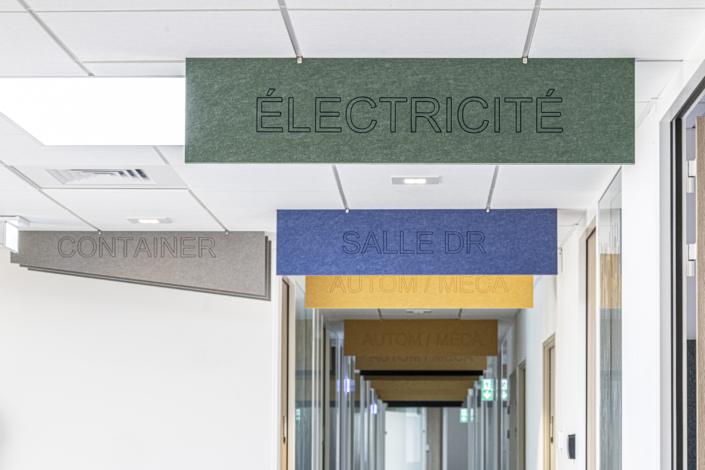 Cartelli colorati appesi al soffitto di un corridoio, con le scritte "Elettricità", "Studio medico" e "Contenitore", creano un'atmosfera dinamica che ricorda una palestra acustica.