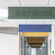 Cartelli colorati appesi al soffitto di un corridoio, con le scritte "Elettricità", "Studio medico" e "Contenitore", creano un'atmosfera dinamica che ricorda una palestra acustica.