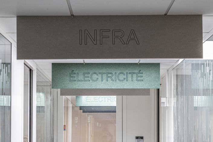 Insegne con le scritte "INFRA" e "ELECTRICITY" sono sospese al soffitto di una moderna sala con pareti in vetro, a testimonianza della cura dedicata all'acustica del palazzetto dello sport, che contribuisce al suo design elegante e funzionale.