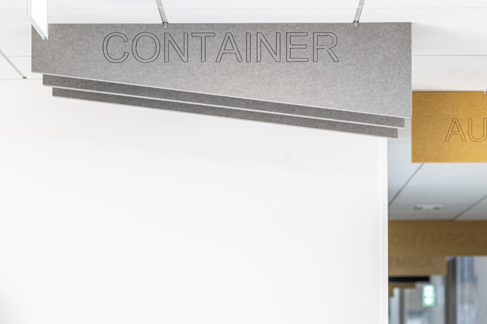Sopra una parete bianca è sospesa un'insegna grigia fissata al soffitto, sulla quale si legge "CONTENITORE"; si intravede anche parte di un'altra insegna, che evoca l'eleganza funzionale tipica del design delle palestre "acustiche".