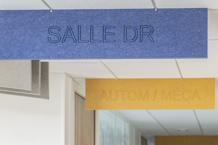 Un cartello blu indica "STANZA DR", e un cartello giallo posto dietro di esso reca la scritta "AUTOM / MECA", in un corridoio con controsoffitto a pannelli bianchi, caratteristico di una palestra acustica.