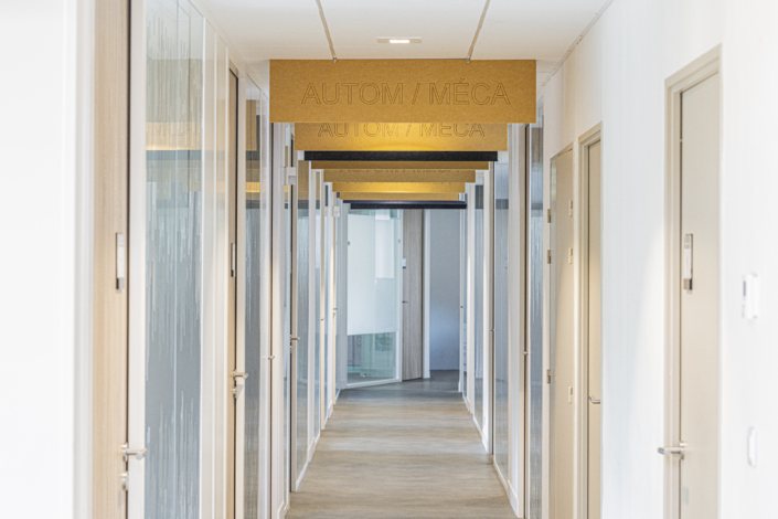 Un moderno atrio d'ingresso con porte a vetri e un'insegna in legno con la scritta "AUTOM/MÉCA" sospesa al soffitto, progettato secondo i principi acustici delle palestre per garantire una buona chiarezza del suono e un comfort ottimale in tutto lo spazio.