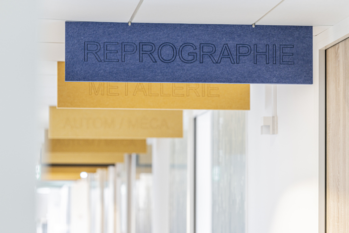 Un'insegna blu con la scritta "REPROGRAPHY" pende dal soffitto di un corridoio, creando un'atmosfera acustica da palestra, con insegne gialle visibili sullo sfondo.